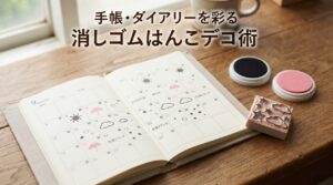 手帳のマンスリーページに消しゴムはんこの枠スタンプ・天気アイコンスタンプが複数色で押され、脇に彫刻済みの消しゴムはんこと無地のスタンプ台が置かれている様子